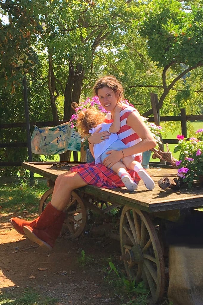 Mamma casual ed elegante con in braccio la figlia su un carretto di fiori, immerse nella natura. Un'immagine di divertimento e coraggio da mamma